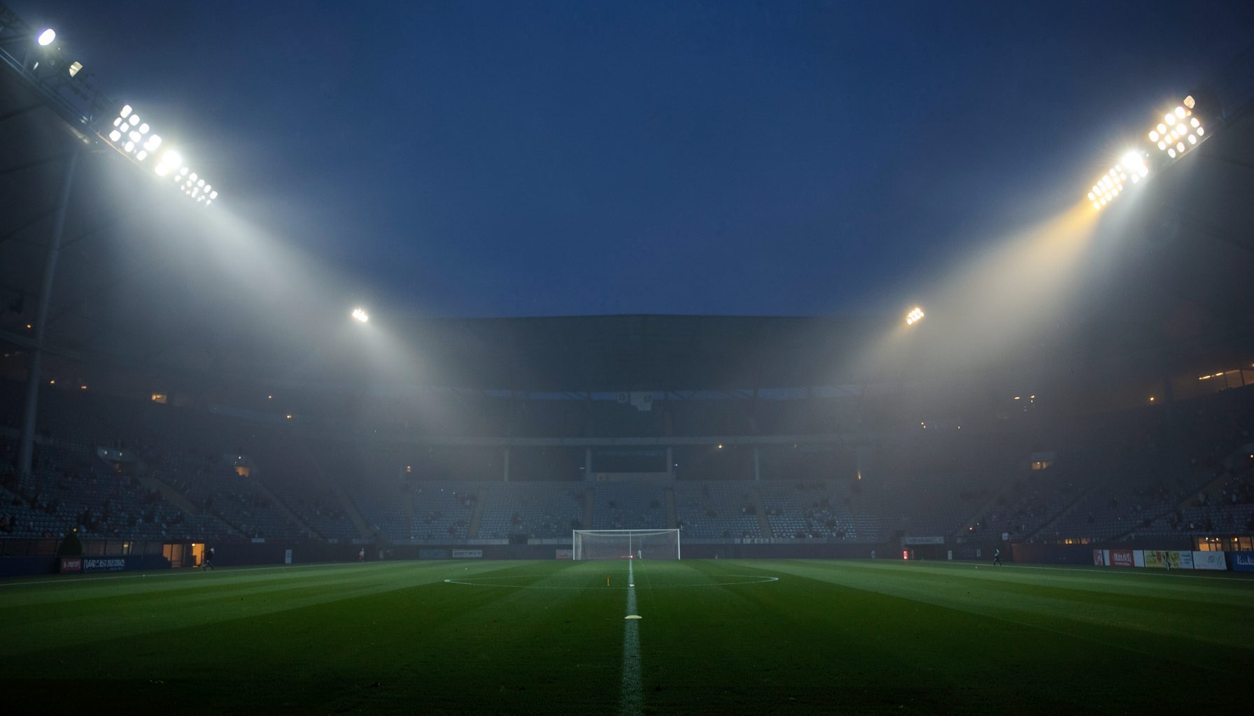 Stadium floodlights piercing through evening mist over a deep green pitch, cool navy and amber tones